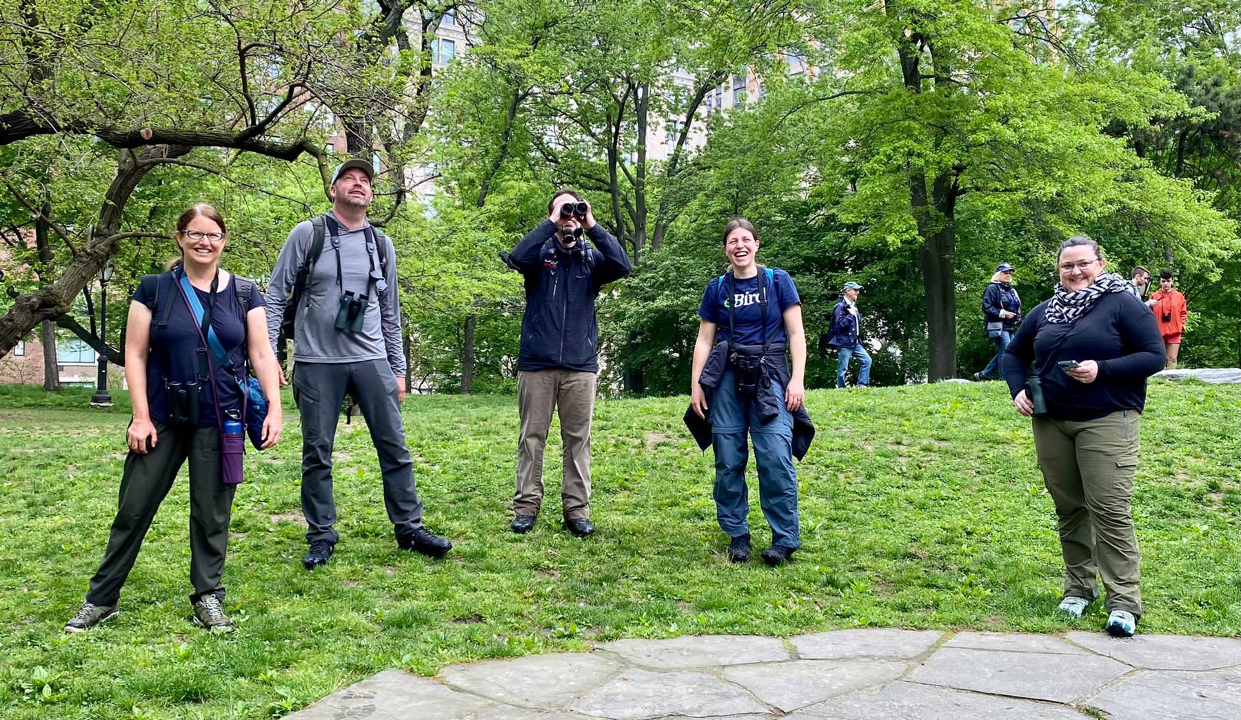five people wearing binoculars, watching birds in a park