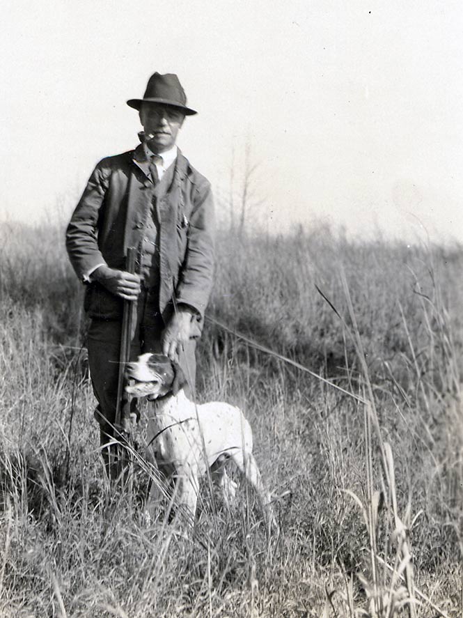 Historical black-and-white photo of a man posing in a field, holding a shotgun, wearing a hunting jacket, hat, and tie, with a hunting dog.