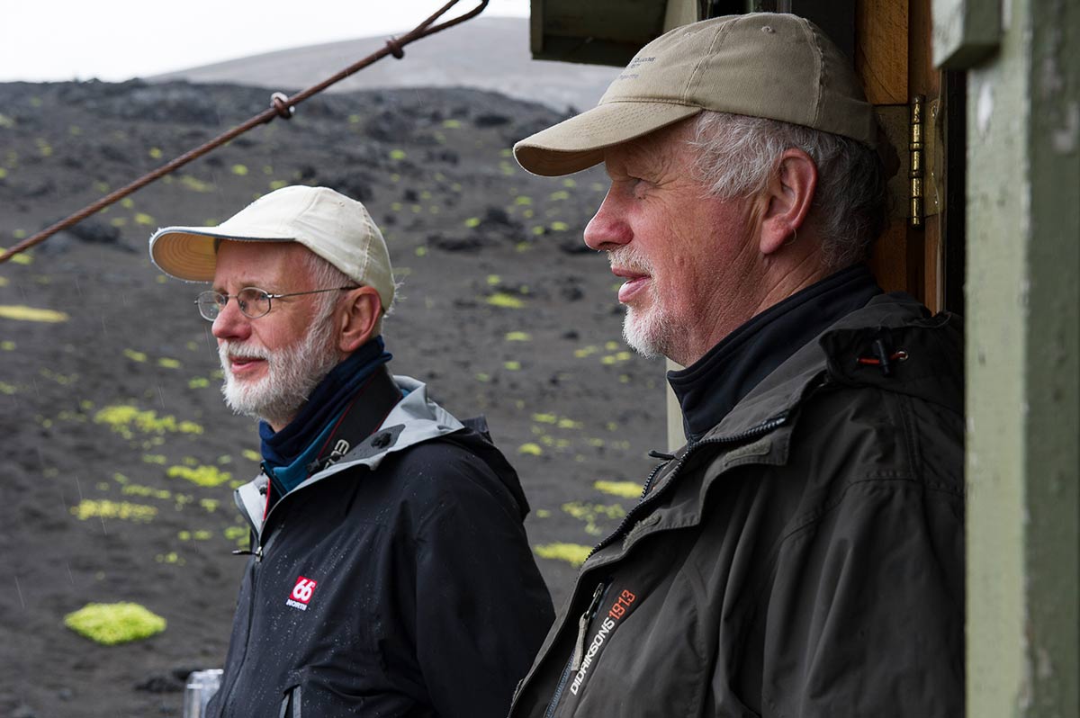 tap for larger version: image of two Icelandic researchers standing outside their research hut with black volcanic ground behind them.