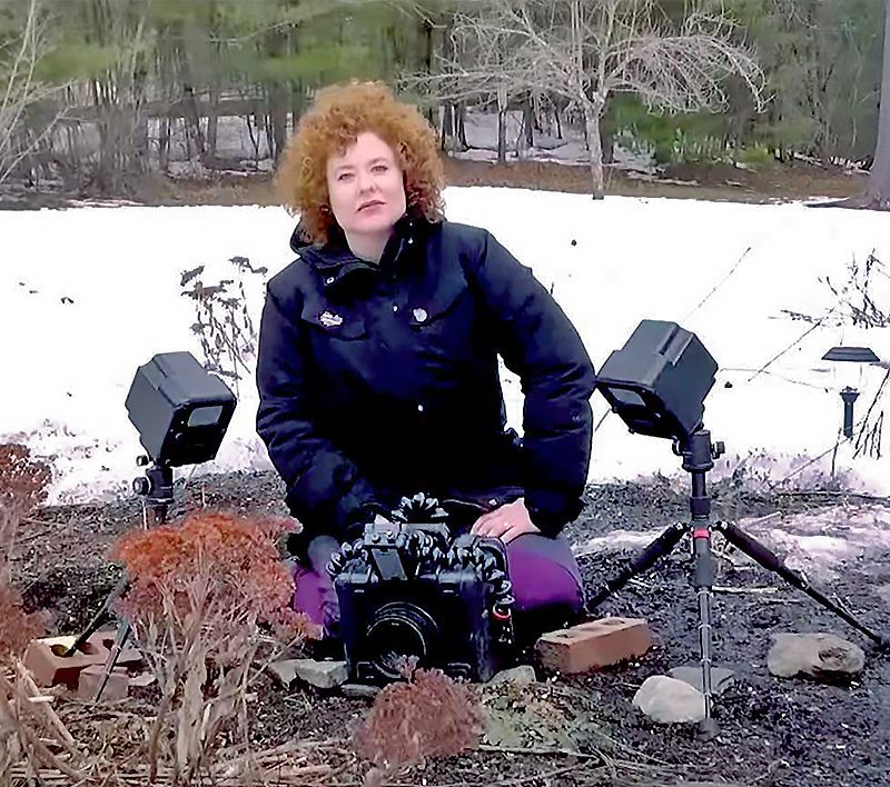 Дама със синьо палто и къдрава червеникава коса седи на земята в снежен ден, заобиколена от оборудване за снимане.