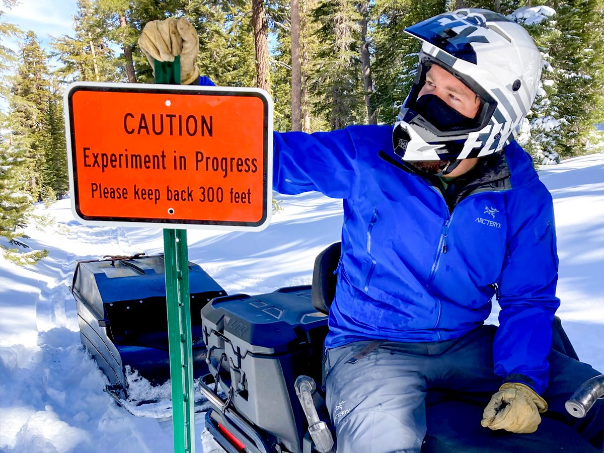 a researcher on a snowmobile in the mountains, next to a sign that reads Caution: Experiment in Progress. Please keep back 300 feet.