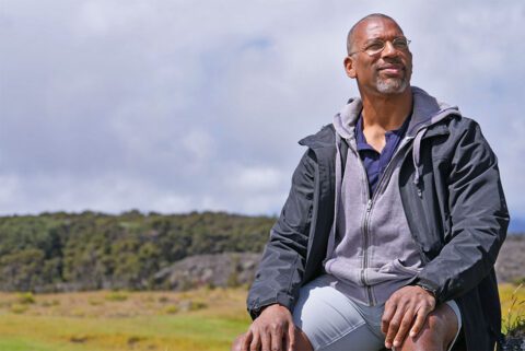 An African American man sits on a rock with hillside vista behind him.