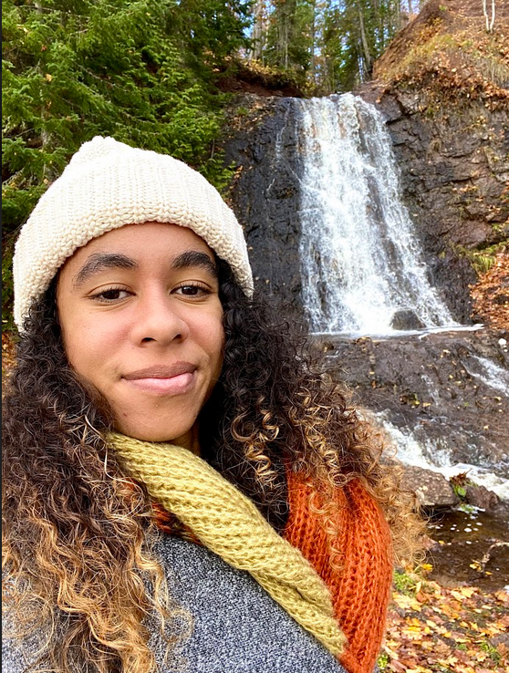 Girl with curly hair and hat stands in front of a waterfall.